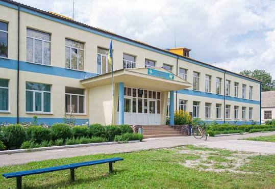 Двоповерхова школа з жовтими стінами, синім оздобленням, прапором і лавкою